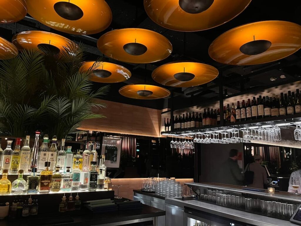 SiSi Glasgow Bar with flat orange lights that resemble disks with an array of drinks at the bar, some people are drinking and chatting in the background.